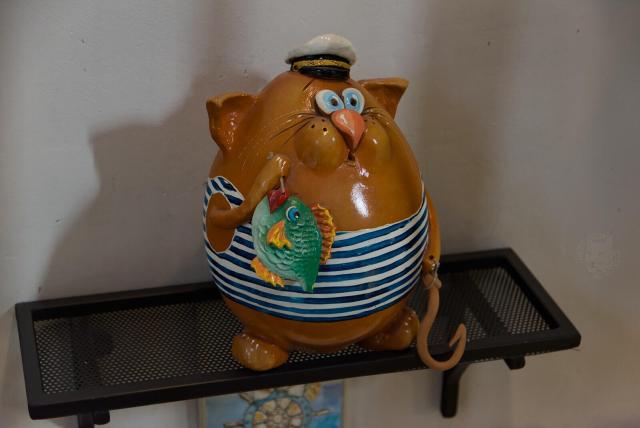 The image features a ceramic figurine of an anthropomorphic cat in a sailor's outfit, complete with a sailor hat. This particular piece is crafted in hues of brown and beige with distinct blue and white striped patterns on its torso, evoking maritime attire reminiscent of sailors' uniforms. The cat is depicted holding a fish in its right paw, and a big fisher's hook in its left paw.

This ceramic figurine stands upright on a black metal shelf against a white wall. The backdrop reveals what appears to be the edge of another decorative item with blue and yellow tones.