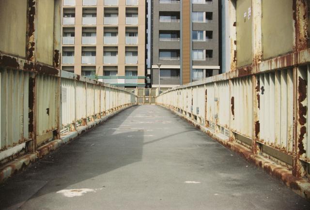The image shows a pedestrian bridge with a metal railing on both sides. The bridge appears to be in an urban setting, with a tall residential building visible in the background. The bridge has a worn and slightly rusted appearance, with visible signs of aging and wear. The surface of the bridge is smooth and paved, and the railing is painted in a light color, possibly white or light yellow, with some areas showing signs of rust and discoloration. The lighting suggests it is daytime, and the shadows indicate the presence of sunlight. The overall scene gives a sense of an older, possibly abandoned or less frequently used urban structure.