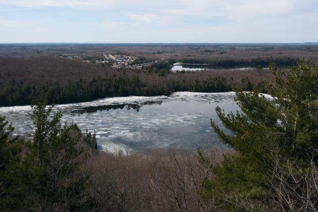 View from a mountain over a lake and a small town beyond in the forest. The ice on the lake is thinning, and a line of trees along the shore is mirrored in an open section of water.