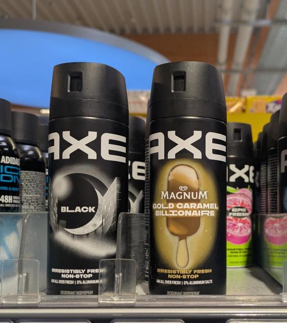 Two cans of Axe deodorant bodyspray sit on a store shelf. The can on the left is "Axe Black" with a silver crescent moon and cityscape graphic. The can on the right is a limited edition "Axe Magnum Gold Caramel Billionaire" featuring an illustration of a Magnum ice cream bar on a stick. Both cans are black with white "AXE" branding.