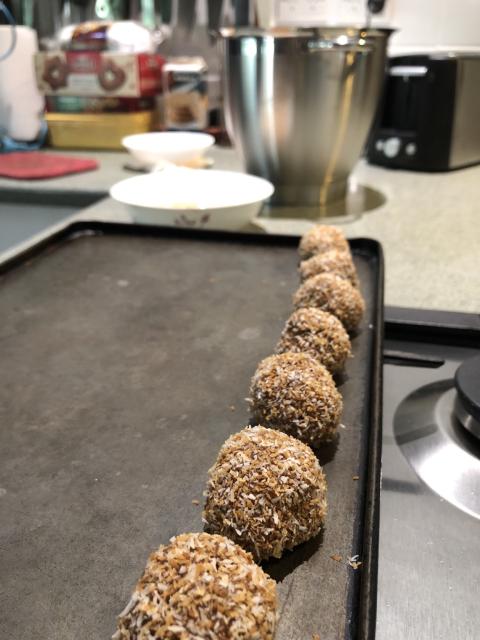 A single row of rum balls on a baking tray. Bowl of toasted coconut and a large mixing bowl in the background.