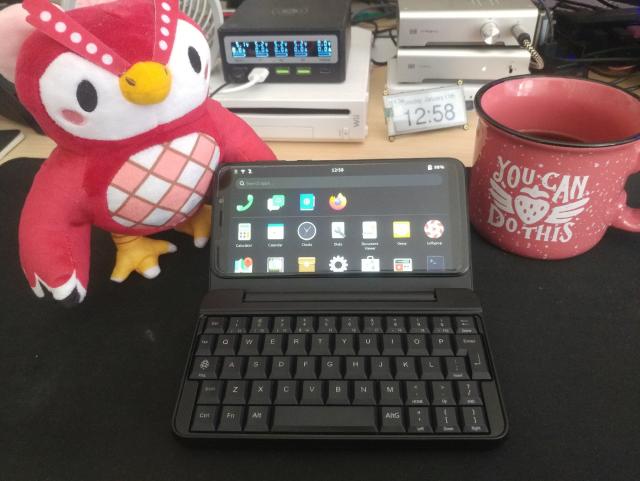 The PinePhone keyboard, opened, next to a plushie of Celeste from Animal Crossing and a coffee mug.