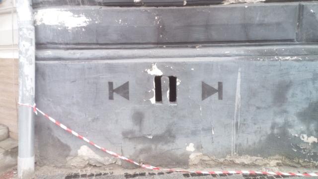 A gray outer wall of a building. In the middle two small holes in of tall rectangular shape, akin to a "pause" symbol. To the left and right, painted two "previous track" and "next track" button symbols.