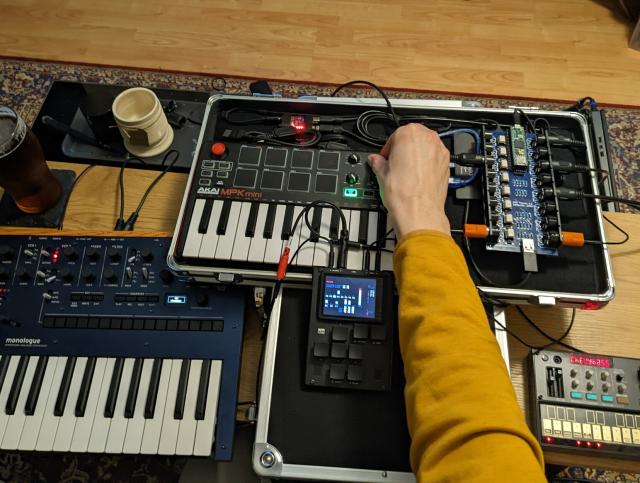 Photograph of a hand tweaking the knob of a MIDI controller connected to a variety of synths.