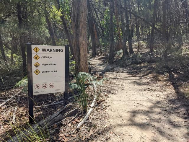 A warning sign next to a bush trail advising of cliff edges, slippery rocks and danger to children. 