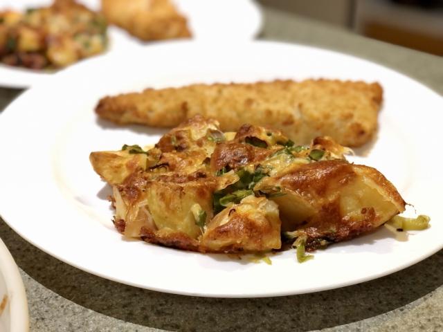 Potato baked with shallots, bacon and cheese, and a crumbed fish fillet in the background.