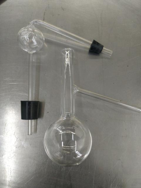 Two pieces of glassware. One is a long necked round bottom flask with a thin off-shoot from the neck slanted downwards. The other is a stoppered thin straw that balloons into a a small bulb where a thin offshoot begins in the bulb and slants downwards. This offshoot also has a stopper.