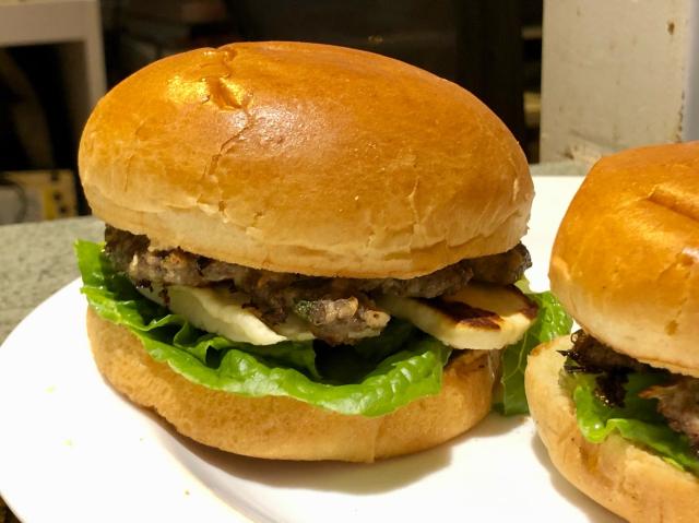 Burger on a plate with another beside it. Can see lettuce, halloumi and lamb patty between the brioche bun halves. 