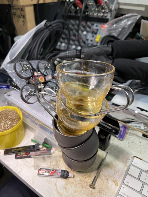 A stack of demitasse, the lower three are elegant: dark brown Japanese sake cups, the top two of which are very Modern Italian glass ones with steel handles. Behind them are a broken micro drone, a pair of shotgun mics taped together. And a mixing board. Scattered around can be seen improvised soldering equipment, a solderless breadboard powered by my proprietary synthesizer daisy chain, and a 12 channel mixing board.