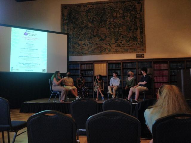 Students sit on the stage by the program projected on the screen. They surround a reporter and are engaged in discussion.
