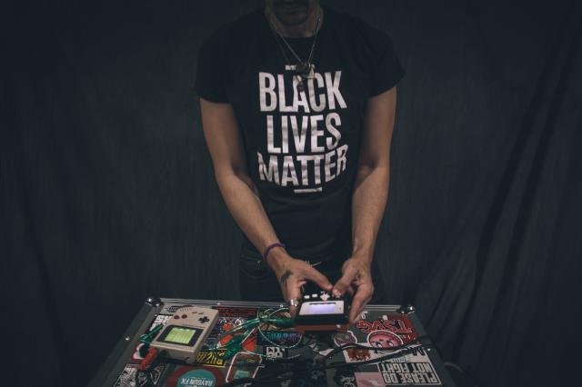 Photo of a person (cropped so their head is not visible) wearing a shirt that reads BLACK LIVES MATTER and making music using two Nintendo Gameboys.