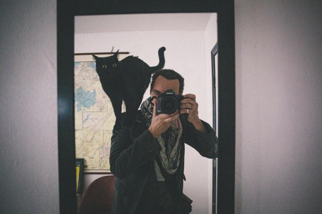 Photo of a photographer taking a photo of themselves in a mirror. Standing on their shoulder is a black cat.