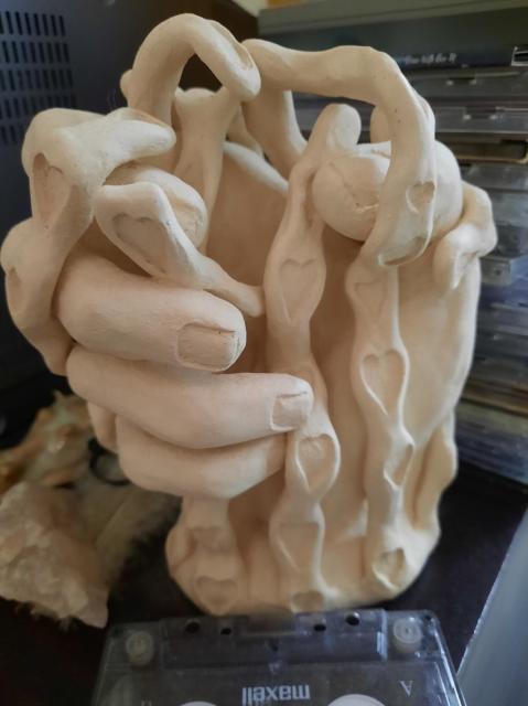 Unglazed ceramic sculpture of a hand, fingers curled around an invisible object and with ribbon-like strands curled around the hand.  The strands have many hearts carved in them.