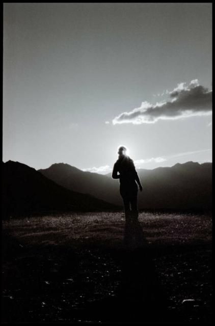 On top of a mountain a woman is standing in front of the sunset and the sun flares like an eclipse.