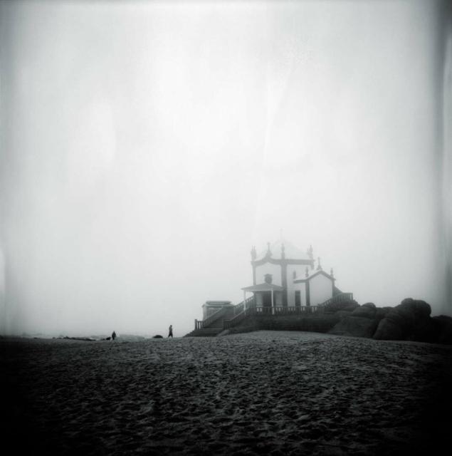 A church on a beach in the fog.
2 persons are walking toward it.