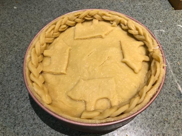 A deep pie filled with sliced roast pork, onion and apple, topped with pastry decorated with 4 pastry pigs and some braiding on the edges. 