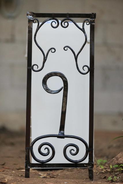 Hand made metal house number panel with scrollwork painted black, standing in front of a cream coloured parcel & letter box. 