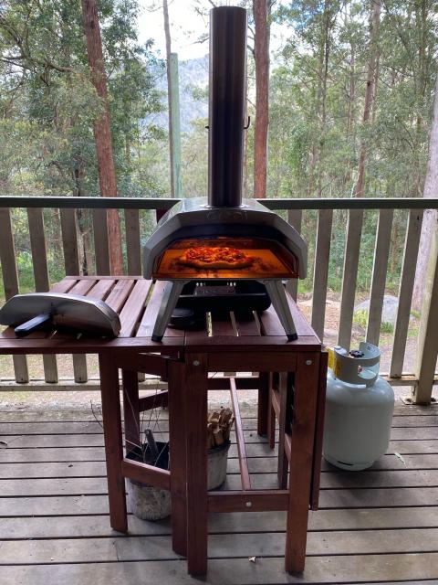 Ooni Karu 12 oven on an Ikea folding table, with a pizza cooking inside. Many trees in the background. 