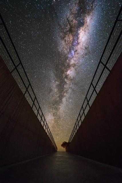 A vertical image showing the Milky Way framed by the walls of a corridor. The Milky Way appears as a diffuse bright band split longitudinally by dark dust lanes. It has subtle orange tones towards the centre, with a few pink/magenta nebulae sprinkled here and there. The Milky Way stretches almost vertically in this image, with its bright core on the upper part. The Milky Way is framed by two high walls of an underground corridor, with hand-rails on top of each wall. At the end of the corridor, where the Milky Way meets the ground, a lone tree can be seen.