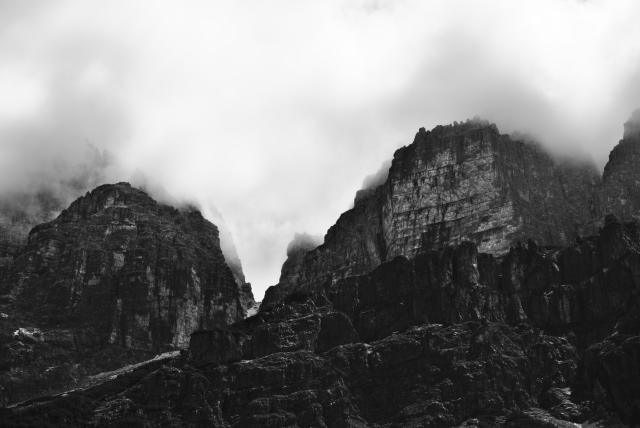 Schwarz-Weiß Bild eines Bergmassivs, aus dem Tal aufgenommen. Die Spitzen der Berge verschwinden in den Wolken. Eine sehr düstere Stimmung.