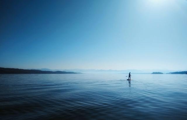 Ein Stand-Up-Paddler auf einem See. Im Hintergrund sind im Diesigen Berge zu erkennen. Es ist strahlender Sonnenschein und das Bild ist in Blautönen gehalten.