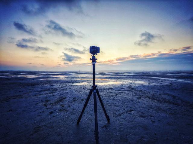 Das gleiche Bild wie vorher nur von hinter der Kamera aufgenommen, sodass eine Kamera auf einem Stativ zu sehen ist. Dahinter der Sonnenuntergang aber mehr in blau und gelb Tönen.