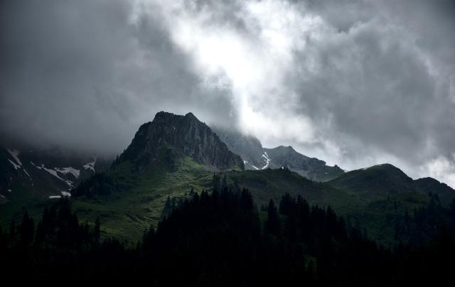 Im Vordergrund sieht man sehr dunkel einige Tannen. Dahinter liegt ein Berggipfel mit einer von der Sonne beleuchteten Gründfläche. Die meisten Teil sind jedoch eher im Schatten. Darüber sind düstere Wolken mit einem Leuchtend hellen Streifen in der Mitte durchden die Grünfläche Beleuchtet wird.