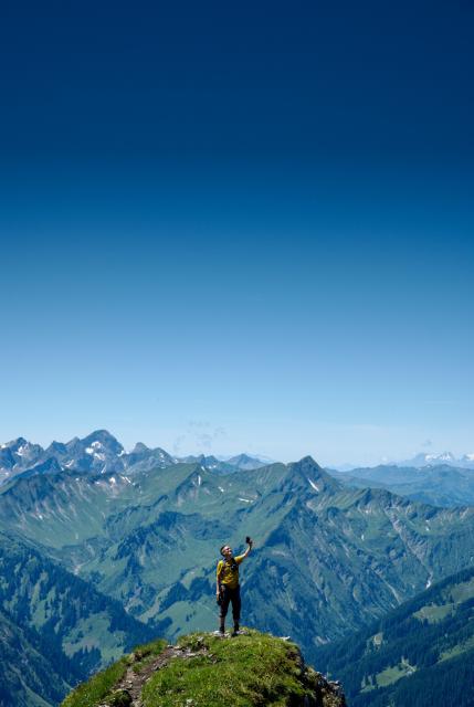 Eine Person mit Rucksacke, gelbem Shirt und schwarzer Hose steht auf einem Felsvorsprung der mit Gras bewachsen ist. Man sieht einen Trampelpfad der zu dieser Stelle führt. Die Person hält ihr Handy hoch um ein Selfie zu machen.
Im Hintergrund sind Berger mit wenigen Bäumen aber doch viel Grün zu erkennen. Einige haben noch ein wenig Schnee auf sich. Der Himmel ist fast wolkenlos blau. 