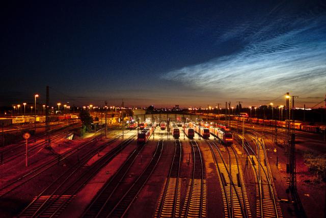 Bild eines Rangierbanhofs mit vielen Laternen. In der Mitte ist eine Wartungshalle und einige Triebwägen zu erkennen die mit einem anderen Licht beleuchtet sind. Der Himme ist dunkel, aber rechts oben sind Nachtleuchtende Wolken zu erkennen.