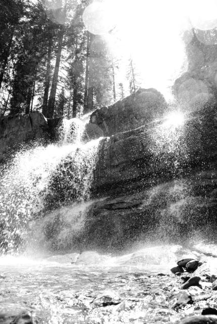 Schwarz-Weiß Bild von einem Wasserfall von unten nach oben aufgenommen. Das Wasser spritzt auf die Kamera zu und läuft dann unter ihr vorbei. Es sind oben links Artefakte von Wassertropen auf dem Objektiv zu erkennen oben Rechts sieht man Nadelbäume stehen.