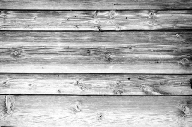 Schwarz-Weiß Bild der Holzfassade an einer Hütte in den Alpen. Man sieht sehr gut die Musterung und an verschiedenen Stellen die Astlöcher.