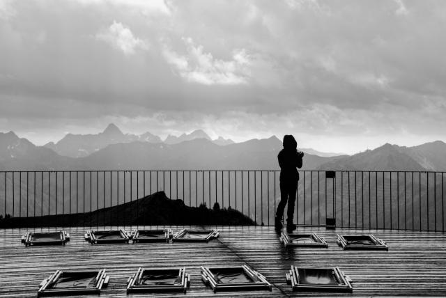 Ein Schwarz-Weiß Bild. Eine Person steht in Regenjacke auf einer Plattform mit nassem glänzendem Holzboden in den Bergen. Sie steht vor einem Geländer und schaut in die Ferne. Auf dem Boden liegen zusammengeklappte Liegestühle und am Himmel sieht man noch die Wolken vom Unwetter.