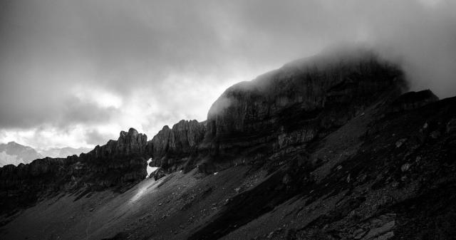 Ein Bergplateau mit einem Geröllfeld darunter. Der Gipfel ist in Wolken gehüllt und ein einzelner Lichtschein erhellt einen Teil des Geröllt. Das Bild ist schwarz weiß
