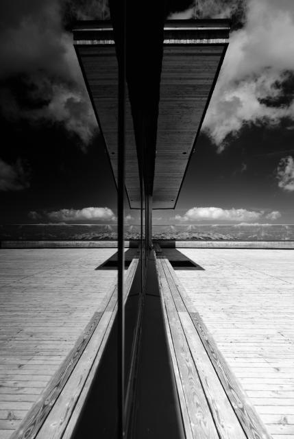 Schwarz-Weiß Bild. Hochkant. Man sieht eine weitestgehend symmetrische Spiegelung einer holz Bank und Terrasse in einer abgedunkelten Scheibe. Ein Teil im Hinteren Bild liegt im Schatten. Mit Mittelgrund ist die Terrasse durch eine Beschriftete Glaswand begrenzt. Der Himmel wirkt dunkel und die Wolken sehr plastisch. Das Bild ist in den Bergen an einer Bergstation einer Seilbahn aufgenommen.