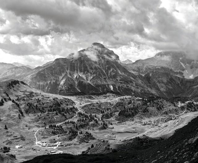 Schwarz-Weiß Bild das ein Alpenpanorama zeigt. Man sieht einige Wege. Bäume in flacherem Gelände, einen See und einen vollständigen Berg. Es ist bewölkt. Das Bild wirkt wie wenn die Vielfalt des Alpenraums in ein Bild komprimiert wurde.