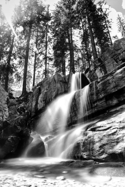 Schwarz-Weiß Bild. Langzeitbelichtung von einem Wasserfall der Dadruch das Wasser als weichen Verlauf anzeigt. Das Wasser fließt über große Felsbrocken in einen kleinen tieferen Bereich. Über dem Wasserfall ragen einige Nadelbäume auf und die Sonne scheint von links Oben ins Bild.
