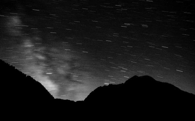 Schwarz-Weiß Bild. Man sieht die Silhouette der Berge ganz in Schwarz. Darüber die vorbeiziehenden Sterne als  Streifen. Links im Bild ist leicht unscharf die Milchstraße zu sehen.