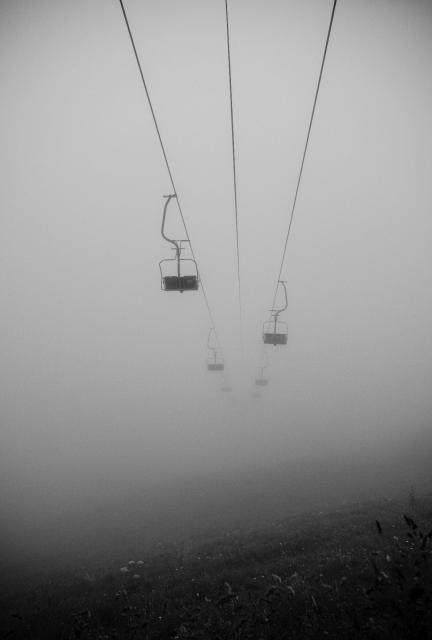 Schwarz-Weiß Bild. Es ist sehr nebelig. Man sieht einen Sessellift im nichts verschwinden. Es ist nicht ganz klar ob er nach oben oder unten fährt. Am unteren Bildrand erknnt man ein wenig Bergwiese die aber auch sehr schnell im Nebel verschwindet. Ein eher düsteres Bild.