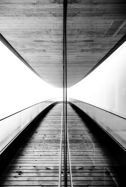 Schwarz-Weiß Bild. Auf dem Bild sieht man eine Terasse aus Holz mit einem Geländer aus Glas. Darüber eine Decke aus Holz. Nach hinten macht das ganze einen Bogen. In der Mitte Reflektiert sich die Szene an einer Glasfront wodurch der Effekt einer Parabel entsteht. An der Galsfront ist noch ein Handlauf aus Stahl angebracht. Im hinteren Teil des Bildes wird es etwas heller. Durch den dichten Nebel ist keine Umgebung erkennbar.