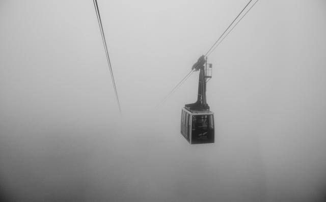 Schwarz-Weiß Bild. Eine Großraumgondel taucht aus dem Nebel auf während die Seile im Nebel verschwinden.