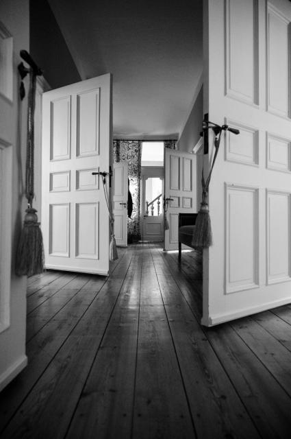 Schwarz-Weiß Bild. Ein Flur mit vielen abgehenden weißen Türen aus denen etwas Licht strömt. Im Fußboden aus Holz spiegelt sich das licht. Im Hintergrund sieht man das Treppenhaus des Hauses.