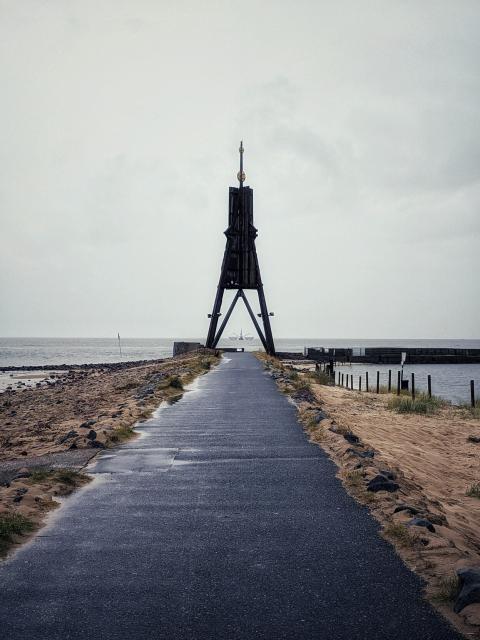 Ein weg führt auf eine große Holzkonstruktion zu. Links und rechts ist Sand und Steine. Im Hintergrund ist Meer. Zwischen den Beinen der Holzkonstruktion sieht man einen Fischkutter mit ausgeklappten Netzen 