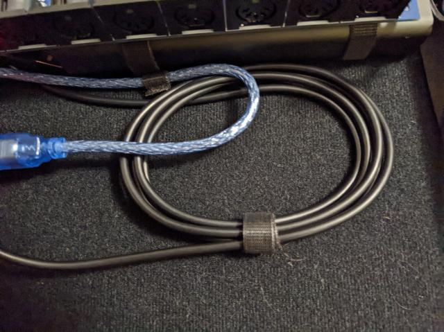 Photograph of keyboard, a couple of MIDI devices, and their respective cables secured down with velcro ties to a base made of the matching velco. Closeup of some cables to illustrate the velco better.