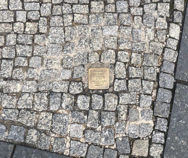 A small square brass plaque set amongst cobblestones in the pavement. It reads Hier Wohnte Dr Georg Lewinsohn  JG 1880 Deportiert 6.3.1943  Ermordet in Auschwitz (Here Lived Dr Georg Lewinsohn born 1880 deported 6.3.1943 Murdered in Auschwitz)