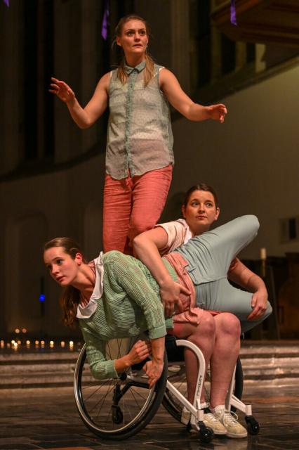 Three dancers on a stage in a church wearing pastel colored clothes.  One lies an the lap of the wheelchair dancer, holding on to the wheel. The wheelchair dancer is holding her. The third dancer is standing on the backrest of the wheelchair.