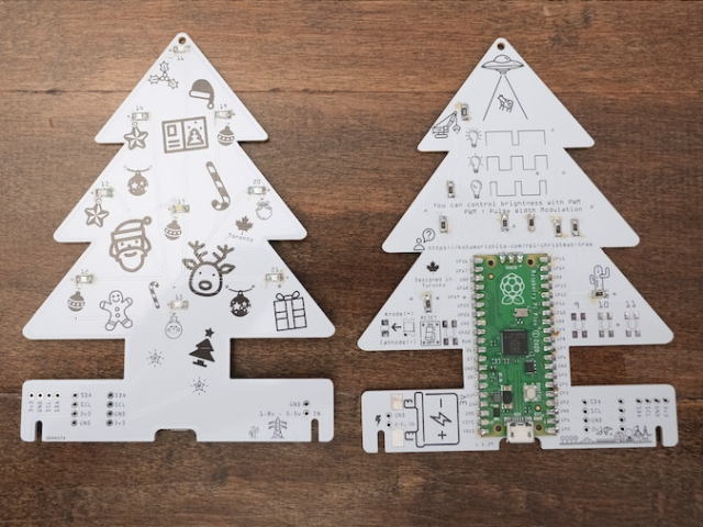 Two white christmas tree shaped P C B boards with black cartoon festive drawings on them lying flat on a wooden table. The board on the right as a Raspberry Pi Pico attached to it.