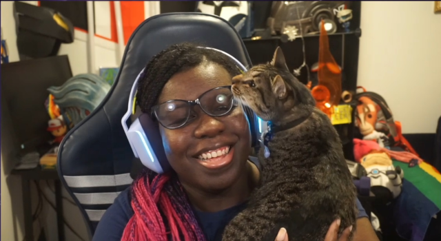 a brown tabby cat being held by a laughing dark-skinned Black person with long black, pink & purple braids, wearing a purple headset and glasses. The cat is rubbing her face against the glasses and knocking them off the person's face. Behind them is a table with shelves full of cosplay props and toys from comic books and video games, including a Hawkgirl helmet, an Asari headpiece, an N7 flag, lightsabers, a Bo-Katan helmet, an N7 helmet, plushies of Mordin, Blasto and Grunt, and a rainbow flag