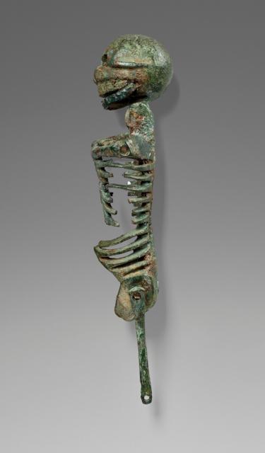Side view of a small bronze articulated skeleton, missing its arms and a leg. It has a green patina throughout