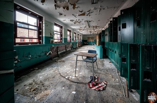 A hallway in an abandoned school. In the center of the foreground there is a desk with attached chair and a crumpled American flag on the floor next to it. Lockers, some open and some closed, line the walls on the right, and windows are on the left wall at periodic intervals. The paint on the ceiling and walls are peeling and the floor is lightly covered with debris