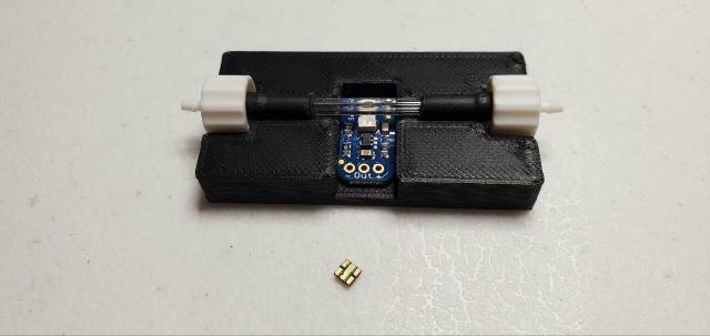 3d printed black jig on a white surface that holds a glass rod, bookended by white luer lock fittings. A small blue pcb sits in a depression in the jig with a small UVC light sensor facing directly into the glass tube. A small gold UVC LED is placed nearby.
